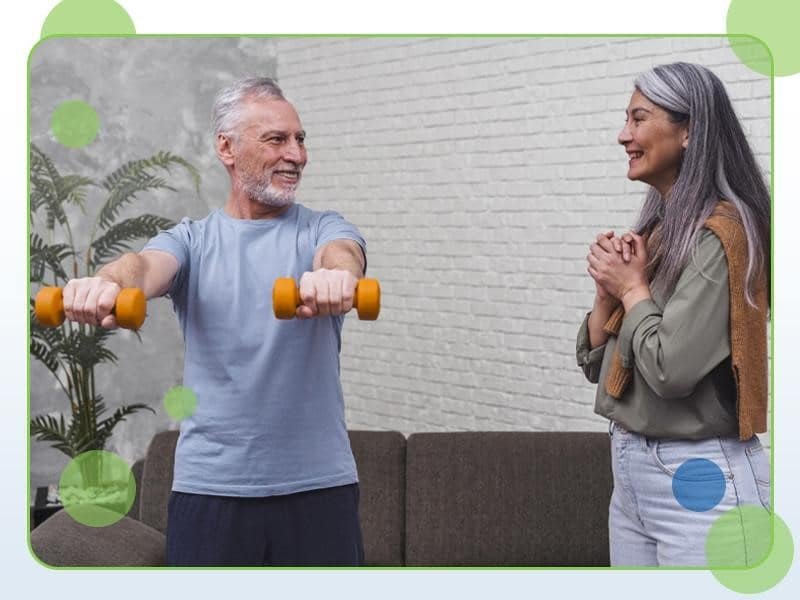 An older man with white hair lifts small orange dumbbells while a smiling woman with long gray hair stands beside him, emphasizing the importance of exercises for seniors. They are indoors, with a couch and a potted plant in the background.