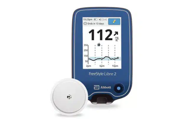 A blue FreeStyle Libre 2 glucose monitoring device from Artik Med displays a reading of 112 mg/dL. A circular sensor is placed next to the device, showcasing the finest technology from the Best Quality Medical Supply store in Florida.