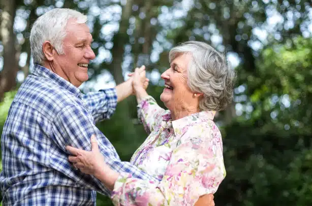 An elderly couple dances outdoors in Florida, smiling and holding hands. They appear happy and joyful, surrounded by greenery and trees, possibly after receiving some care from Artik Med medical supply.