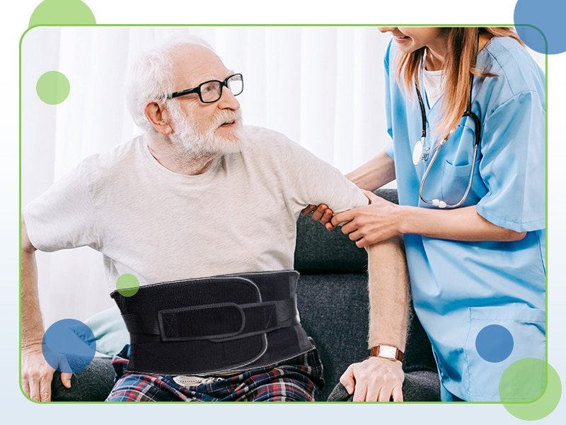 An elderly man wearing glasses and a back brace is assisted by a nurse in a blue uniform. Seated on the couch, he receives support for back pain relief as she helps him stand up.